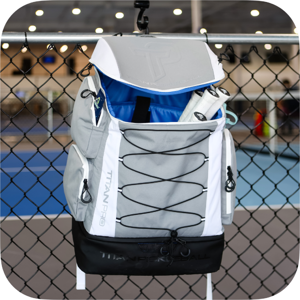 Gray and white Titan Pickleball Backpack with multiple compartments hanging on fence at indoor court