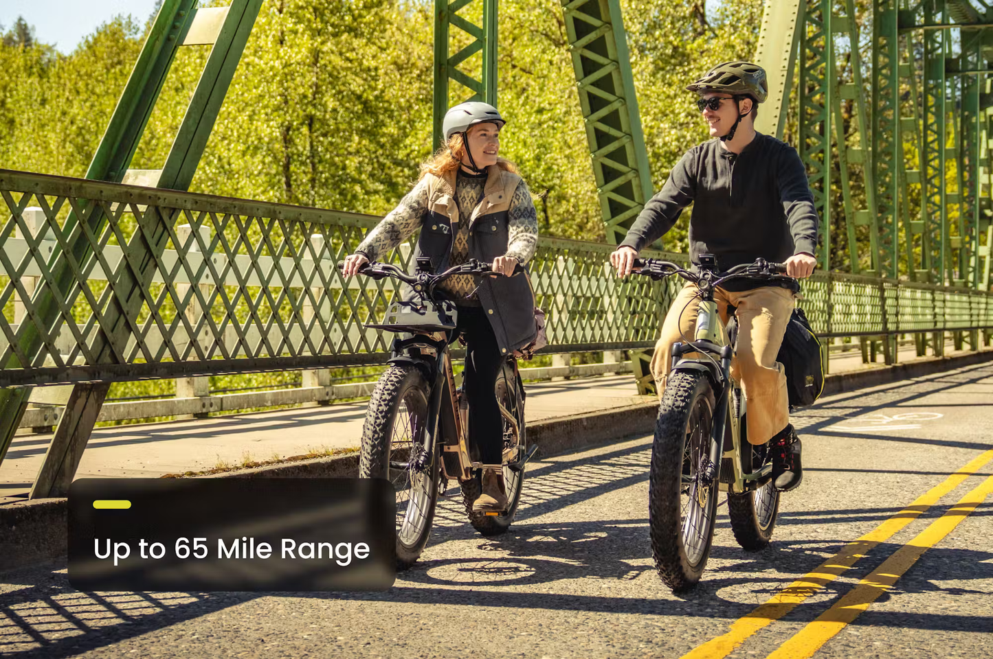 Two people riding Aventon Aventure.3 step-through e-bike by Rex E-Bikes on a sunny bridge with up to 65 mile range