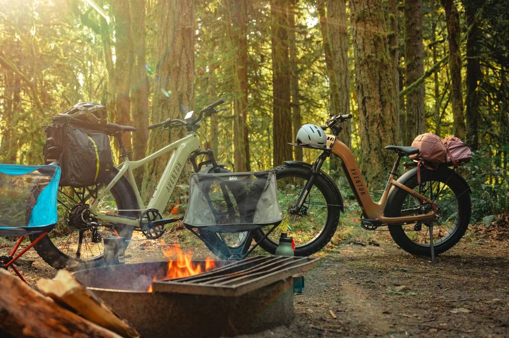 Two Aventon Aventure.3 e-bikes for sale parked near a campfire in a forest setting with backpacks attached