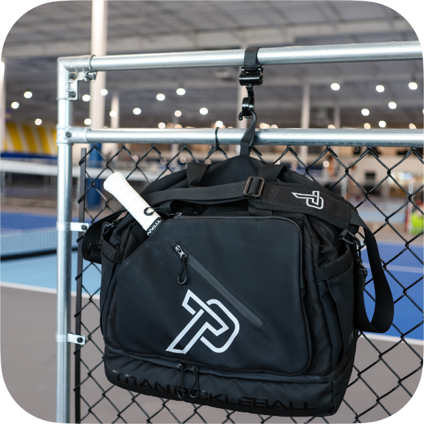 Black Titan duffle bag hanging on chain link fence with pickleball paddle inside at sports court