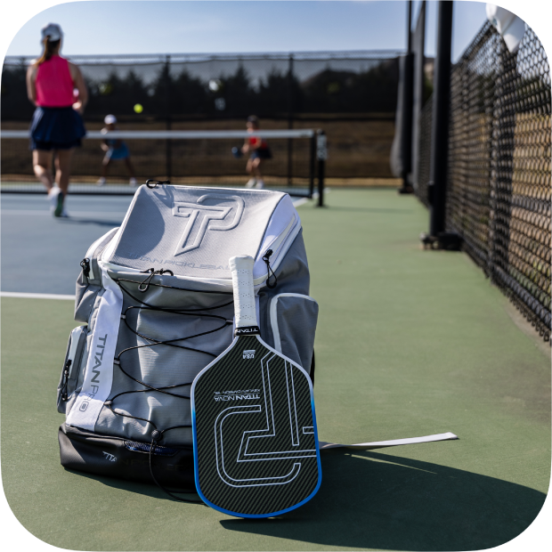 Gray Titan Pickleball Backpack with paddle pocket on outdoor pickleball court with players in background