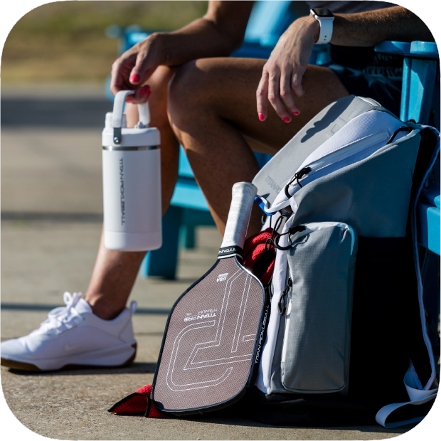 Titan Pro Titanium pickleball paddle by Titan Pickleball leaning against a gray backpack with person holding a water bottle